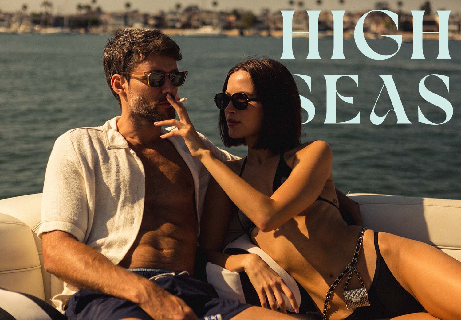 Couple relaxing on a boat in swimwear, woman touching mans face. High Seas text visible in the background.